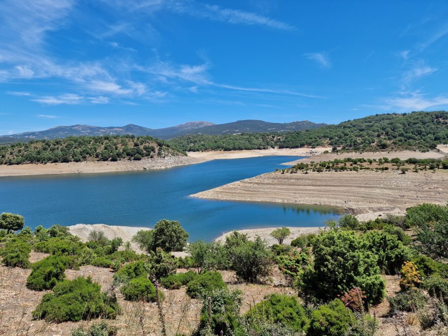 Stille Oase Blick auf einen See umgeben von Hügeln und grüner Vegetation unter blauem Himmel.